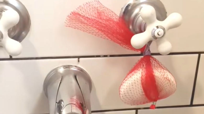 Bar soap wrapped in produce netting over bathtub faucet on white tiles