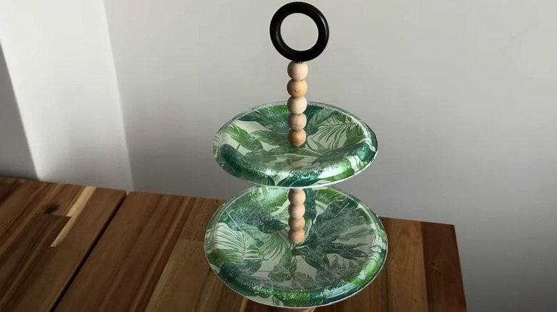 A floral cake stand sitting on a wooden table top in a room corner.