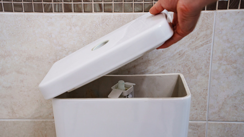 A hand removing the lid of a white porcelain toilet tank