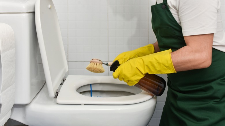 Close-up of hands wearing yellow cleaning gloves scrubbing a toilet with a brush while spraying eco-friendly disinfectant