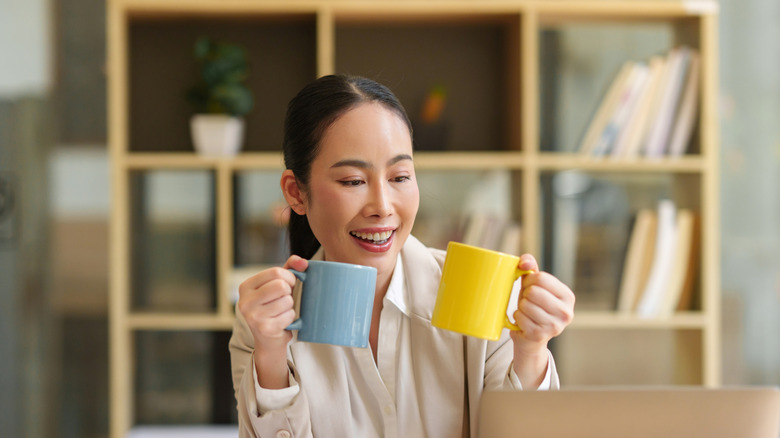 A woman smiles while holding two colorful coffee mugs