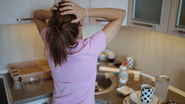 A frustrated woman holds her head while looking at cluttered kitchen countertop