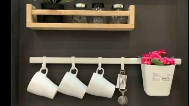 A Sunnersta rail system hangs on a kitchen wall holding three mugs, a tea steeper, and a small flowering plant.