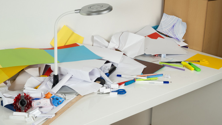 A cluttered desk covered in papers, pens, and a lamp
