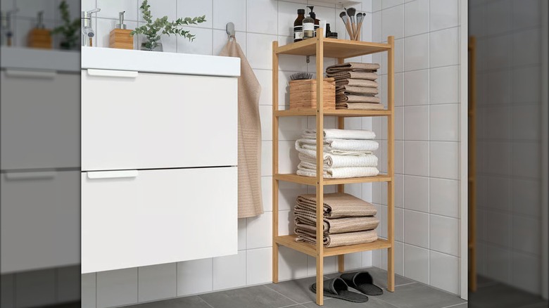 Toiletry items and folded linens are stacked on a bamboo shelving unit in the corner of a white bathroom