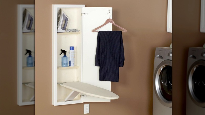 An in-wall ironing board cabinet open with board extended and a pair of pants hanging from the garment hook on the door