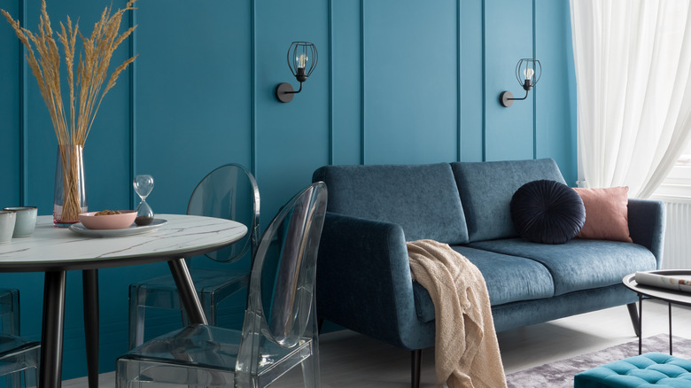 A small living room with teal walls, a small, round glass table, a blue couch, a light purple rug, and white curtains