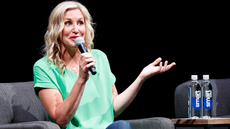 Jenny Marrs holding a microphone and talking to a crowd while sitting on a gray sofa.