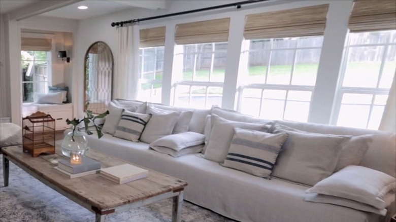 A sunny gray and white living room featuring light wood Roman shades with white airy curtains over the large windows behind the couch