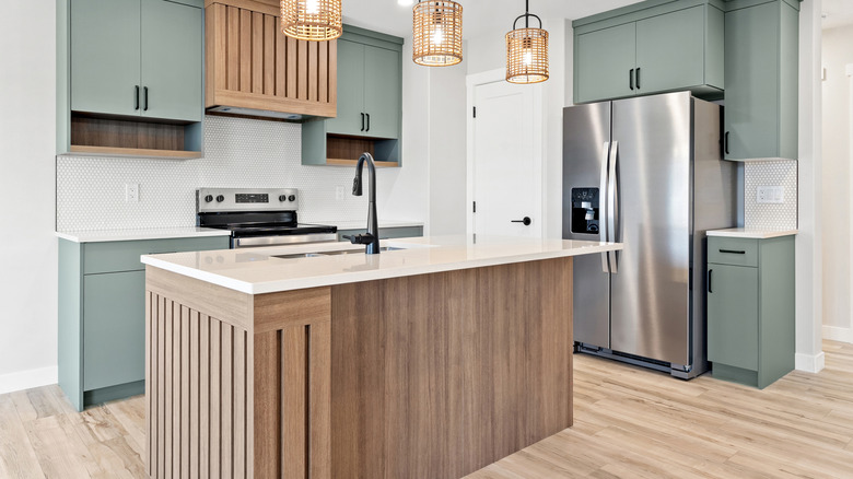 A brown kitchen island with a marble top in a well lit kitchen with green cabinets