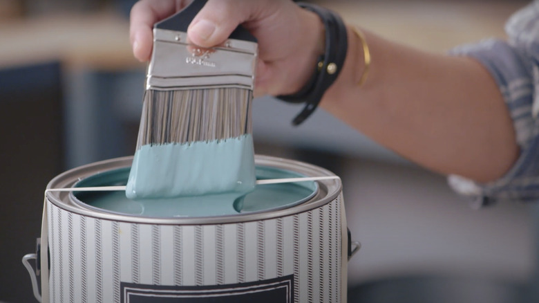 Joanna Gaines wiping excess paint off of a paint brush on a rubber band stretches around an open paint can
