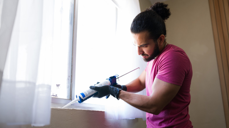 A young man with long hair uses caulk to seal windows