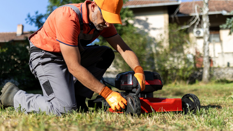 A man kneels as grass sticks to underside of lawn mower