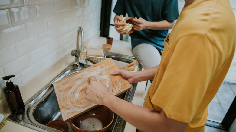 A person scrubs a wooden cutting board over a kitchen sink