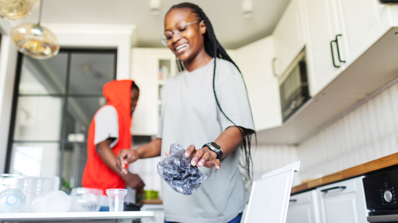 A young Black woman smiles while throwing trash away in a kitchen garbage can