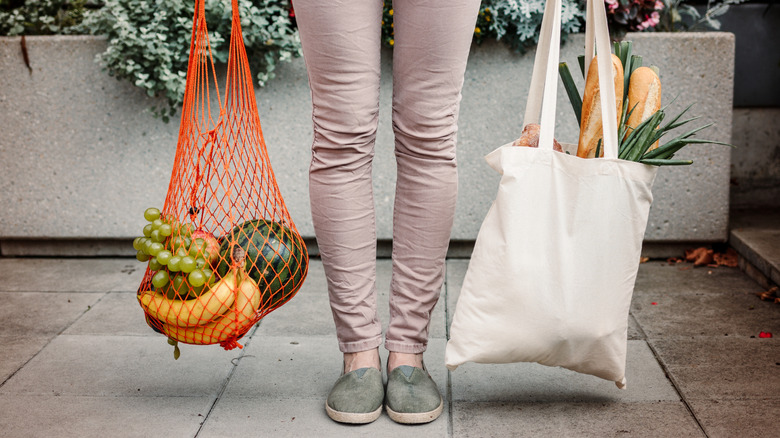 A person holds a reusable white canvas bag and an orange mesh bag full of groceries