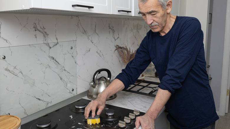An elderly man uses a yellow sponge to clean a stovetop