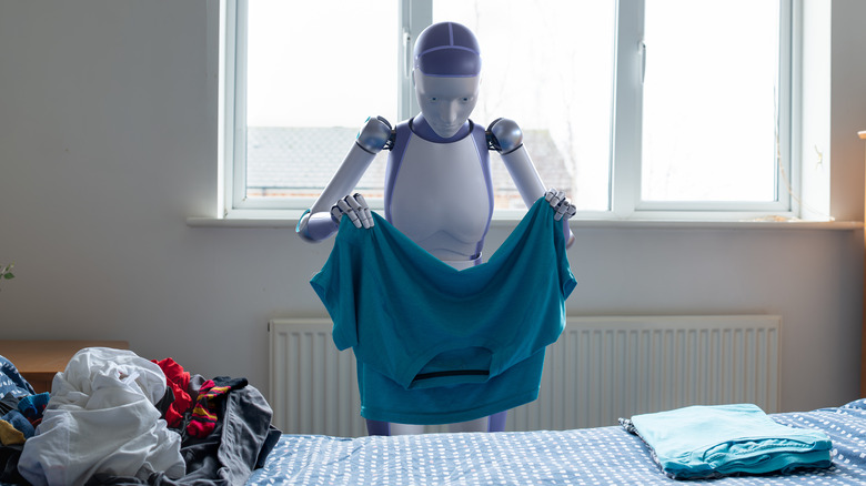 Generic white and purple humanoid android robot is holding up a blue shirt to fold it on the bed