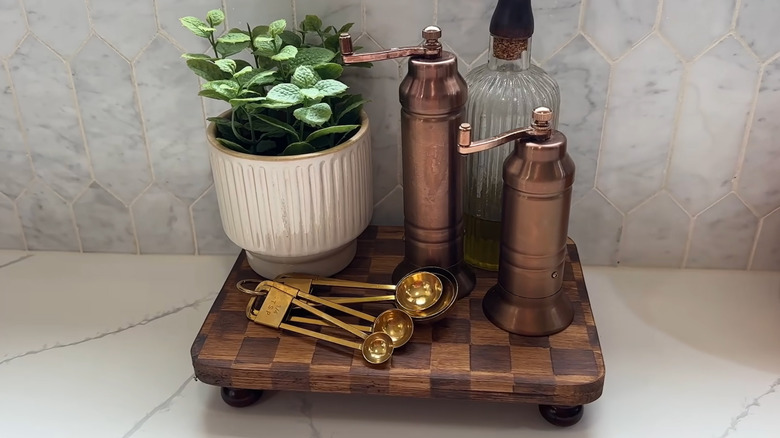 Measuring spoons, salt and pepper grinders, and a potted plant sit on a Magnolia checkered wood riser dupe