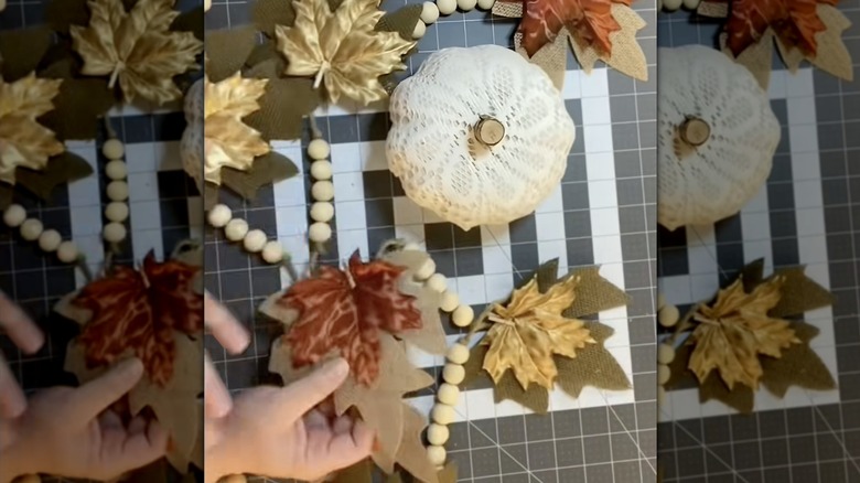 A white pumpkin with a doily next to a fall garland on a crafting table