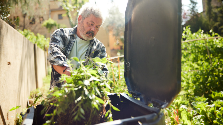 A person dumping weeds into a plastic garbage can near a large wooden privacy fence