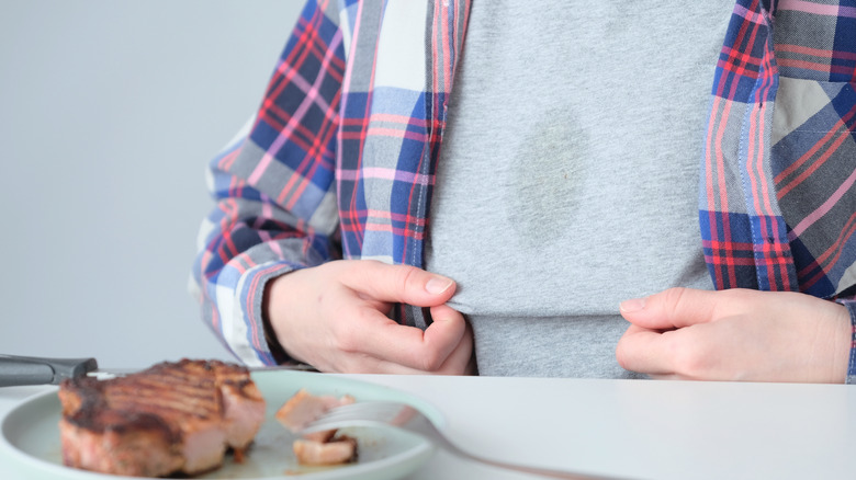 Man sitting at a table in front of a plate of meat withn oil stain on his grey shirt