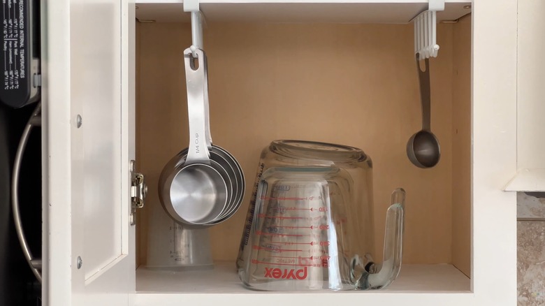Small metal measuring cups hanging on mug rack beside upside-down glass measuring cups