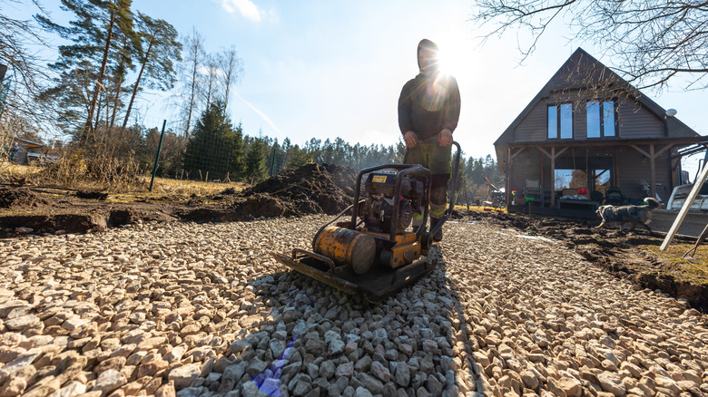 A person compacting a gravel driveway in front of a cottage