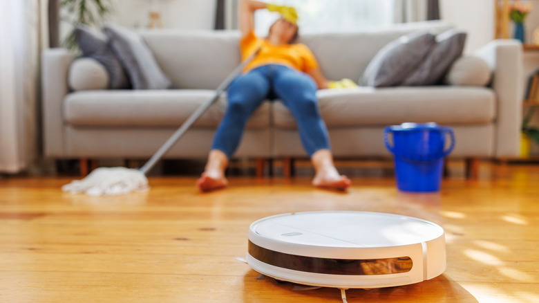 A tired person collapses on a couch while a robot mop gets to work