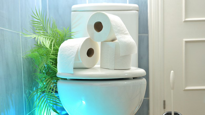 White toilet in blue tile bathroom with three toilet paper rolls stacked on the lid