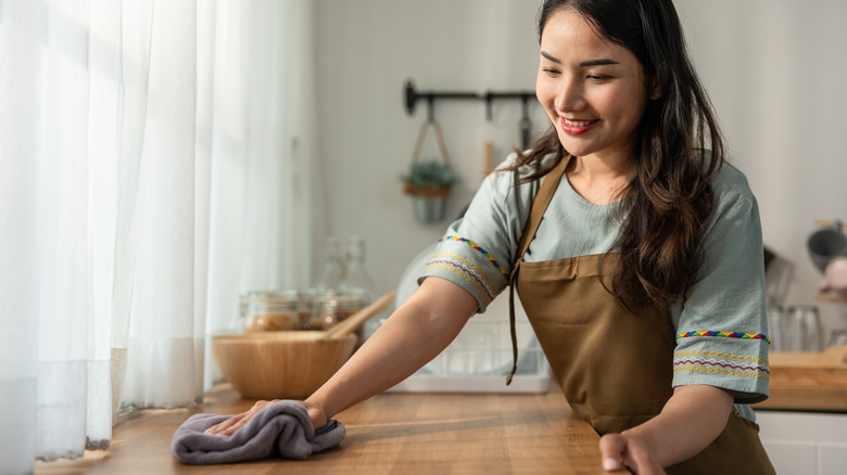 Smiling woman wipes a wooden kitchen countertop with a gray rag