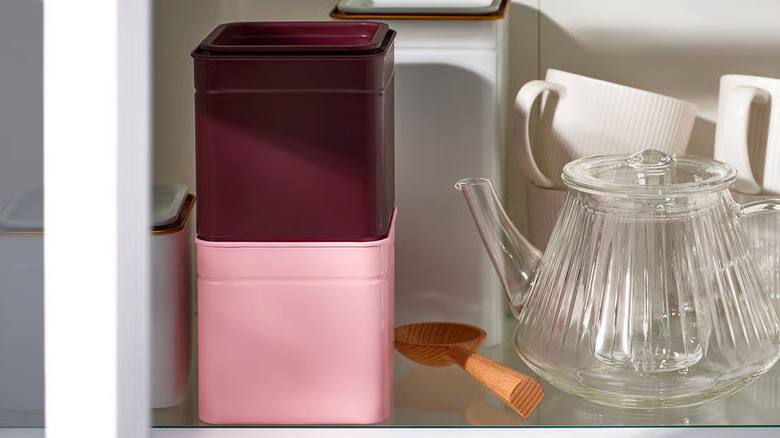 Two colorful metal tea tins sit on a shelf next to a glass teapot