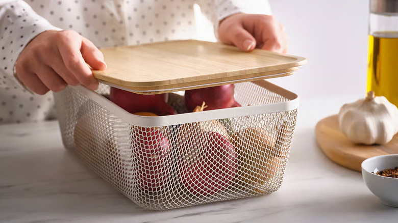 A person's hands are shown putting a wooden top on a wire basket filled with produce