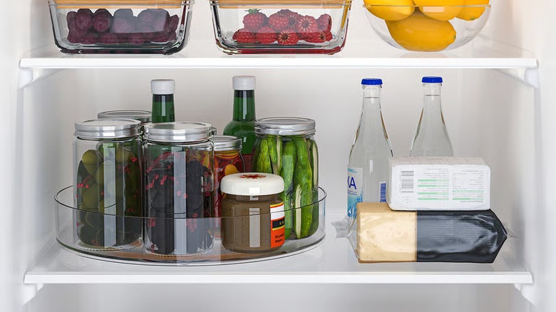 Glass condiment jars sit inside a clear plastic lazy Susan on a refrigerator shelf