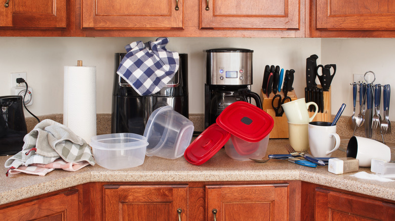 Cluttered kitchen countertop with appliances, mugs, storage containers, and dish towels