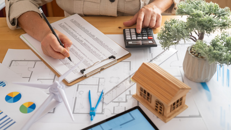someone doing math and looking at a clipboard with a wooden house and other math tools in foreground