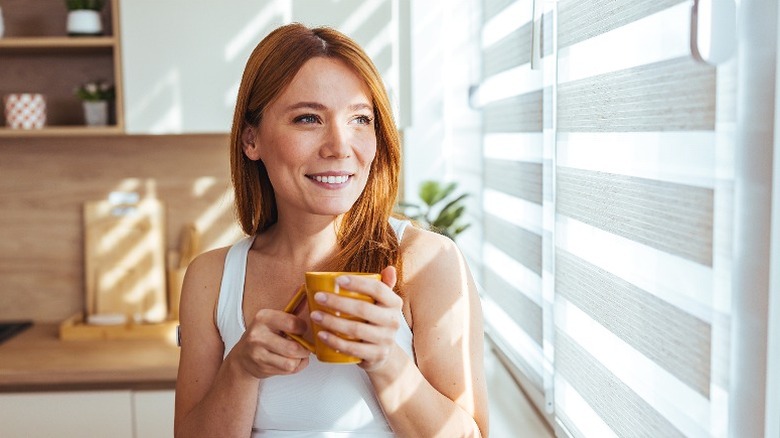 Smiling woman drinking coffee in kitchen while looking through window blinds