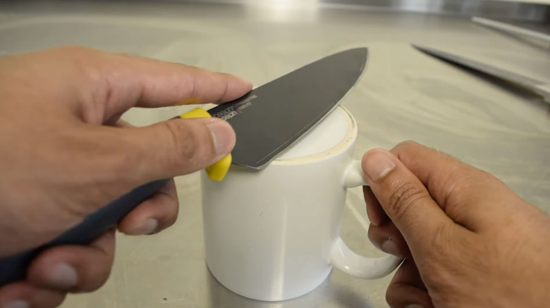 A person runs a large kitchen knife against the unglazed ring of a ceramic mug