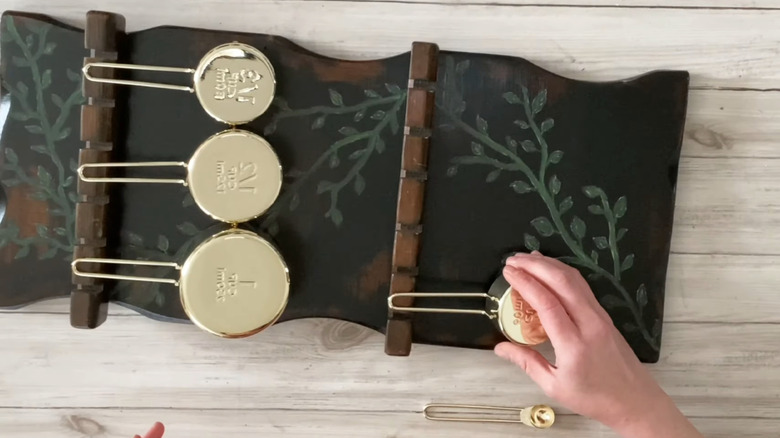 A person upcycles a spoon rack to hold a gold measuring cup set