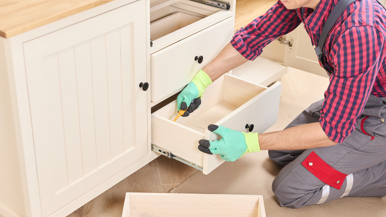 Man with colorful gloves and screwdriver disassembling farmhouse-style drawers from cabinet