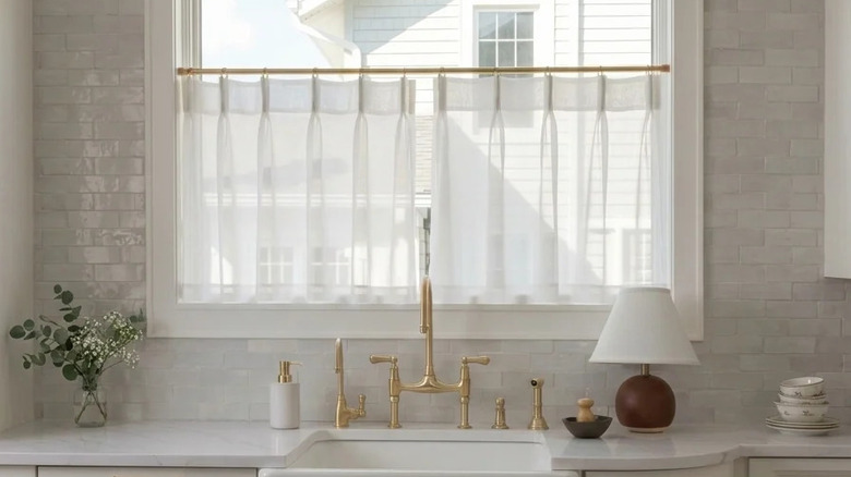 A kitchen with grey tiled walls, a butler sink, and sheer cafe curtains in the window