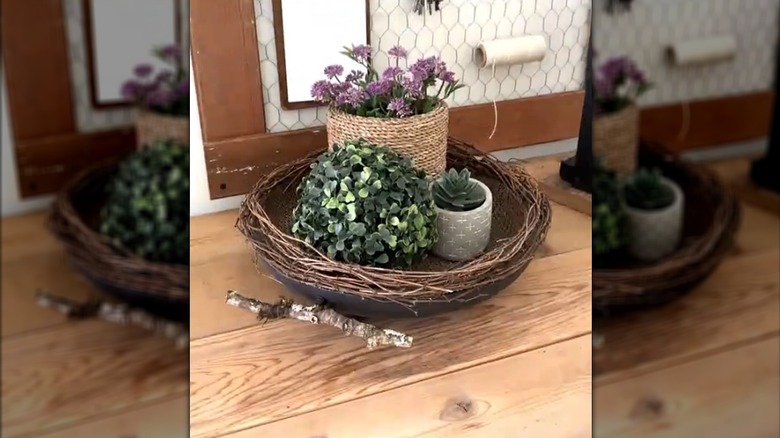 Frying pan lined with twigs and filled with artificial foliage arrangement on wooden outdoor workbench