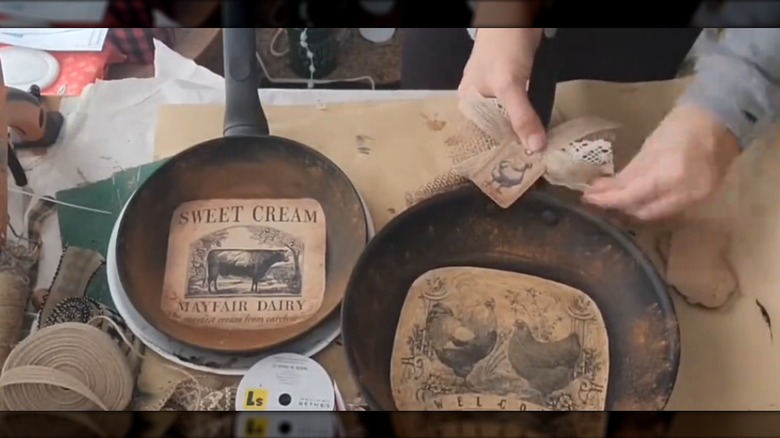 Old frying pans with decoupaged farmhouse images and rustic burlap bows on crafting table