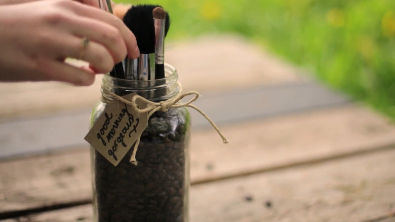 Person arranging makeup brushes in a mason jar filled with coffee beans