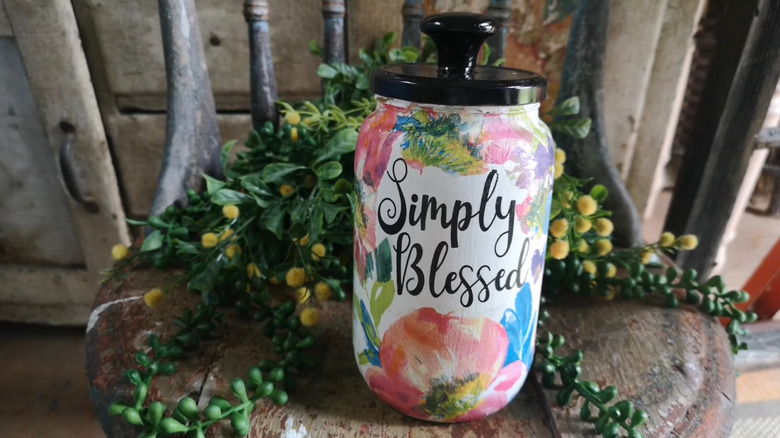 A pickle jar decoupaged with flowers sitting on a small wood table