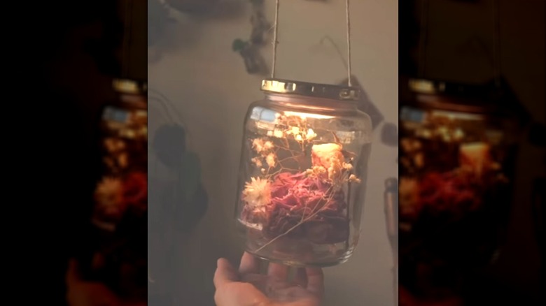 A pickle jar filled with dried flowers and a light on the lid being held by a person's hand