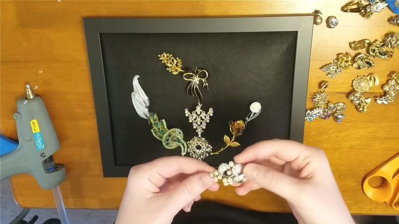 Woman tacking vintage gemstone brooches to a modern black frame with opaque black fabric backing on wood craft table