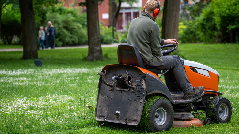 A man mowing the lawn on an orange riding mower while the family looks on in the background.