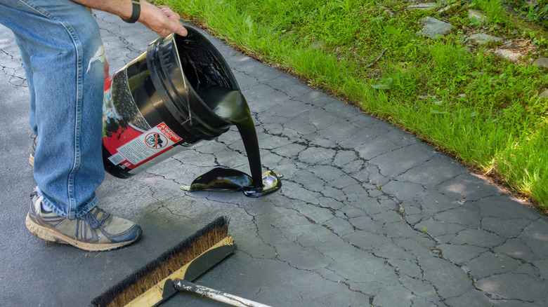 A person pours black liquid from a bucket to repair cracks on an asphalt driveway.