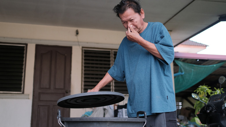 A man holds his nose while standing in front of a smelly trash can